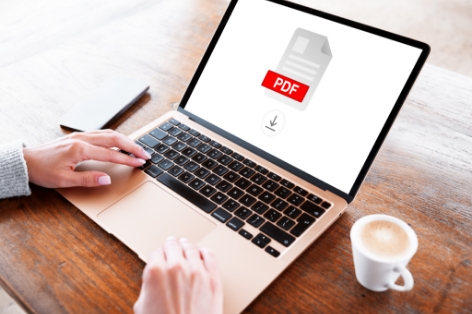 Person typing on a laptop at a wooden table with a PDF icon displayed on the screen and a cup of coffee nearby.