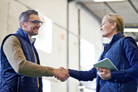 Two professionals shake hands in a bright warehouse; man in glasses wearing a navy vest, tan sweater, woman in blue jacket holds a tablet, both smiling.